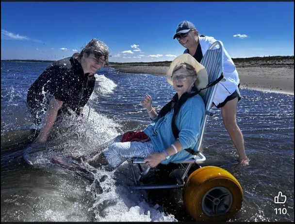 A person using an all-terrain beach wheelchair enjoys the shoreline at Pomquet Beach while two companions assist them through the shallow water. Waves splash around the wheelchair as the group shares a joyful moment outdoors, highlighting accessible recreation and inclusion.